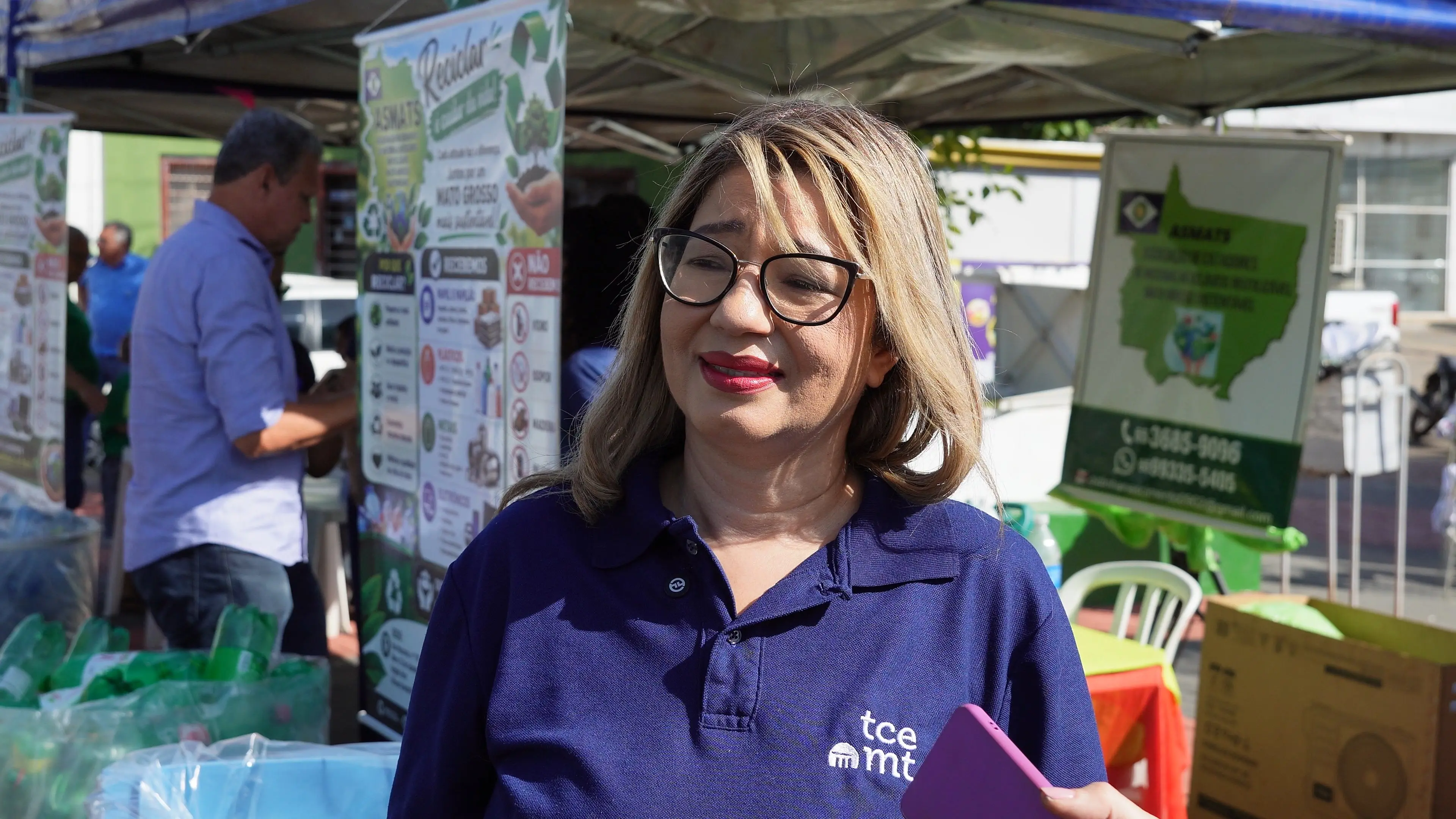Mulher de óculos e camisa azul-marinho do TCE-MT participa de entrevista. Ao fundo, tendas da ação ambiental exibem materiais recicláveis e painéis informativos sobre coleta seletiva.