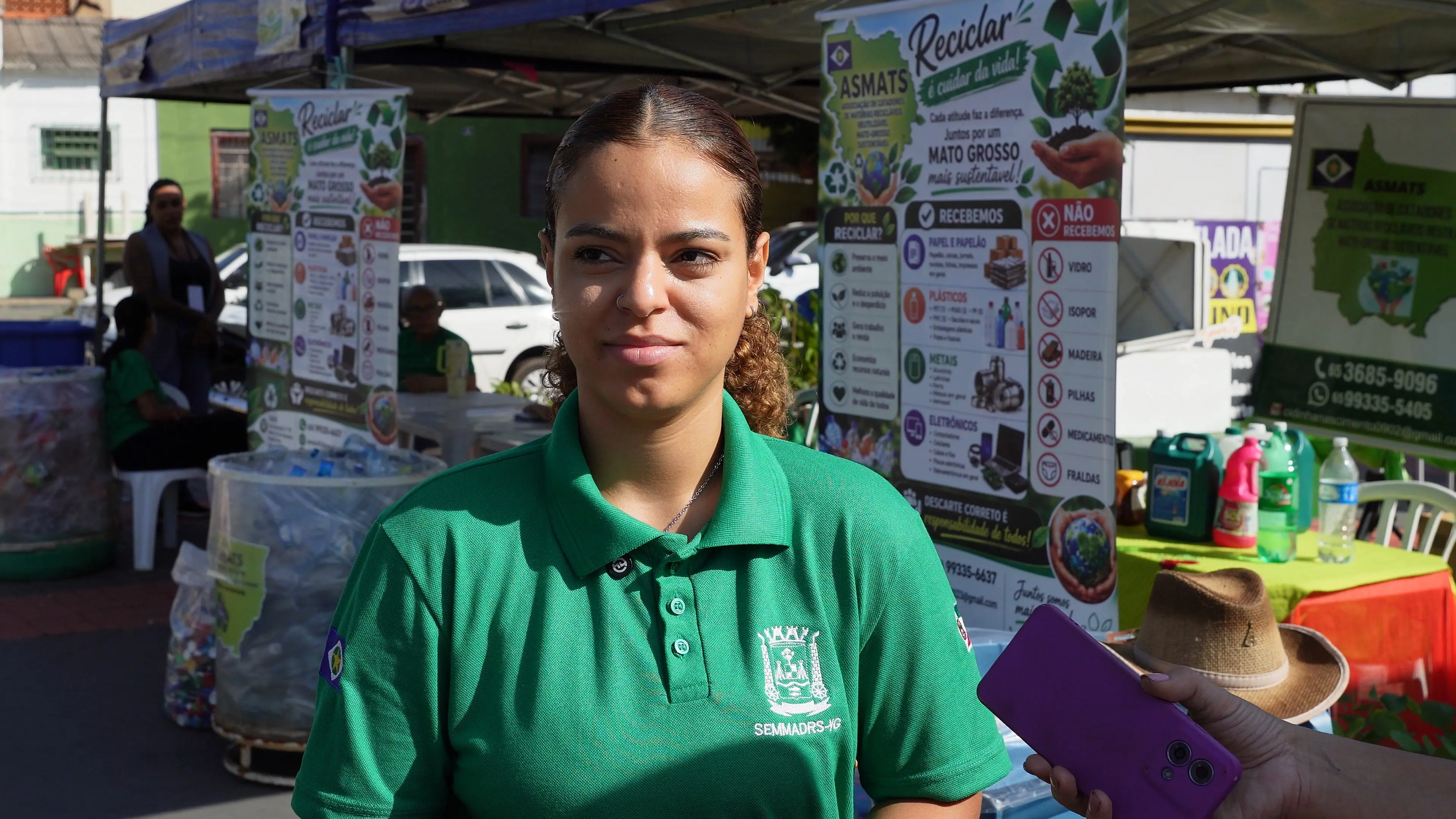 Jovem de camisa verde da Secretaria Municipal de Meio Ambiente de Várzea Grande fala durante entrevista. Atrás dela, banners educativos e materiais recicláveis compõem o cenário da ação.