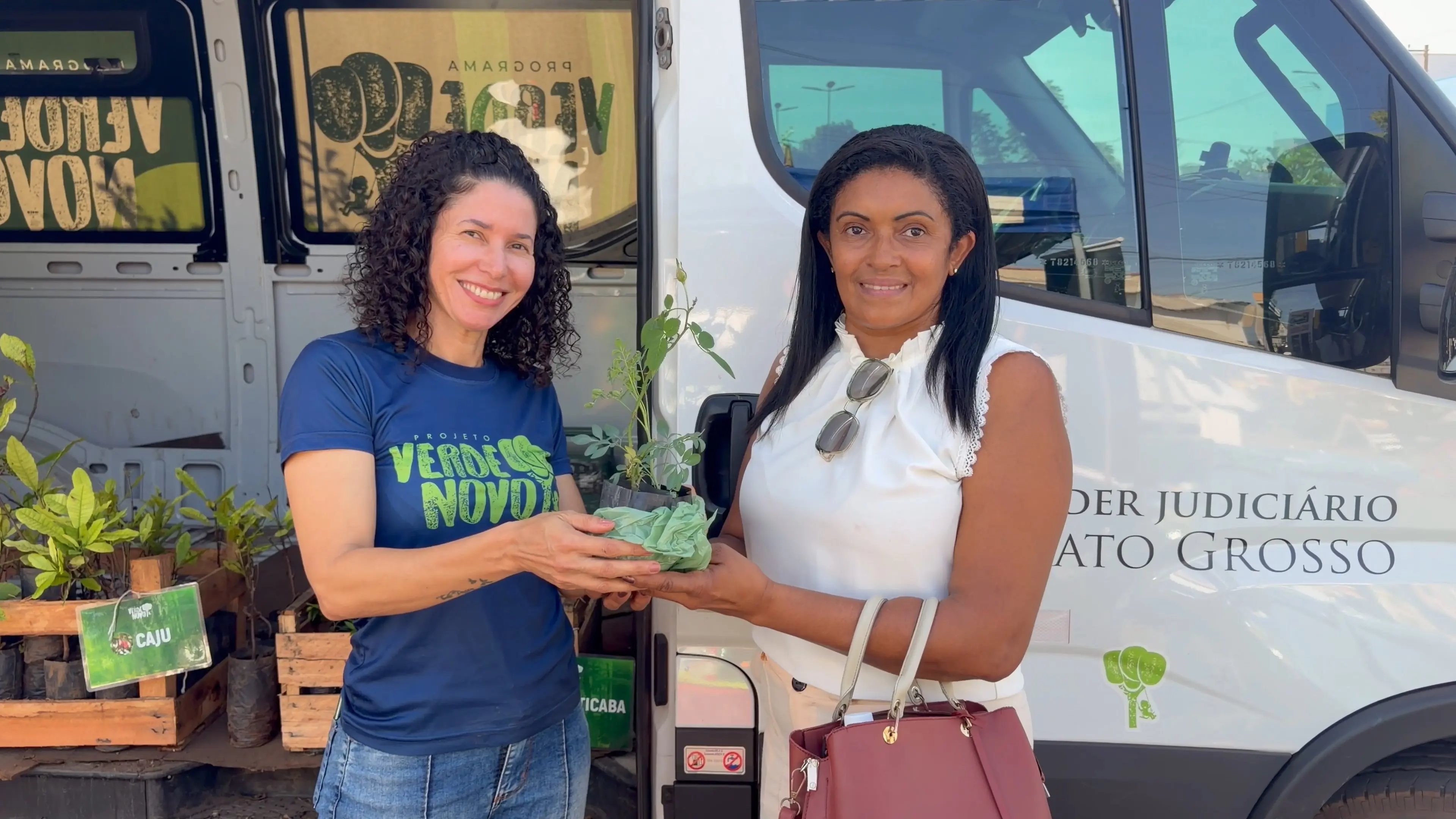 Representante do Programa Verde Novo entrega uma muda de árvore a uma moradora. As duas sorriem diante do veículo do projeto, simbolizando o incentivo à arborização e ao cuidado ambiental.
