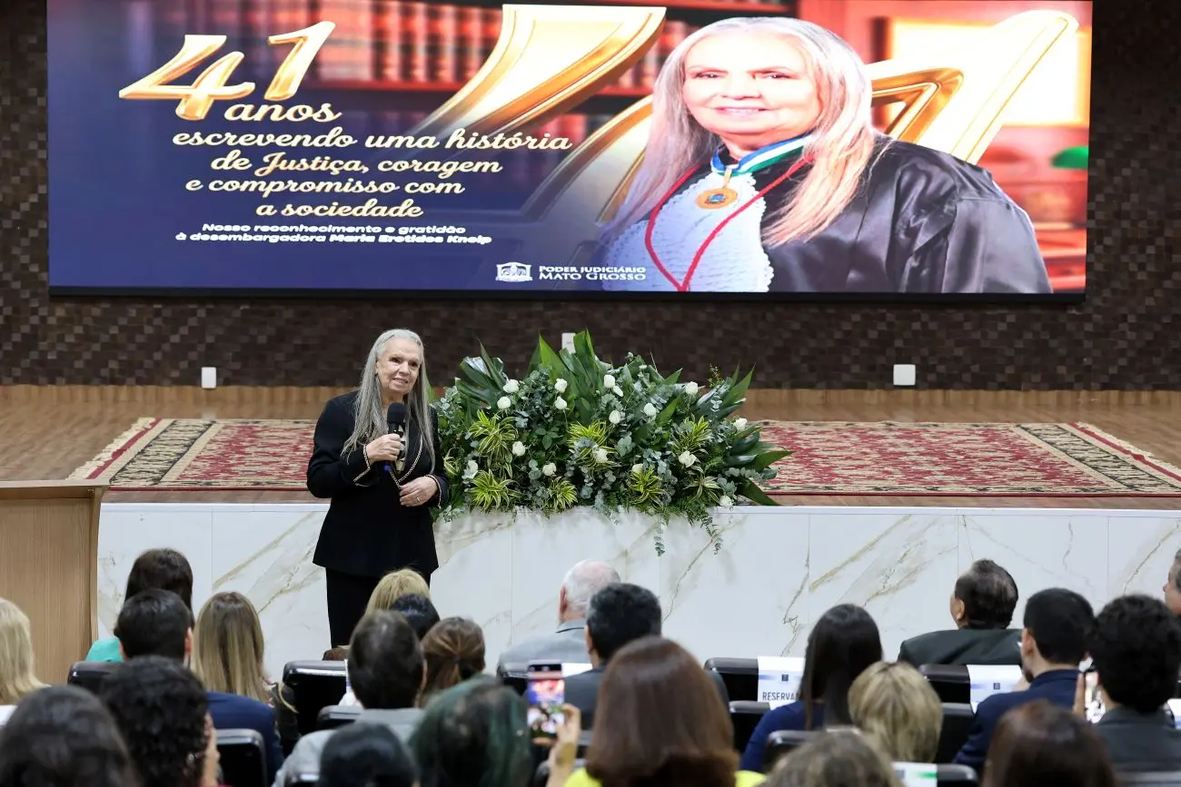 Foto horizontal que mostra a desembargadora Maria Erotides Kneip em pé, falando ao público, no auditório do TJMT. No palco, o telão exibe uma arte em sua homenagem com sua foto.