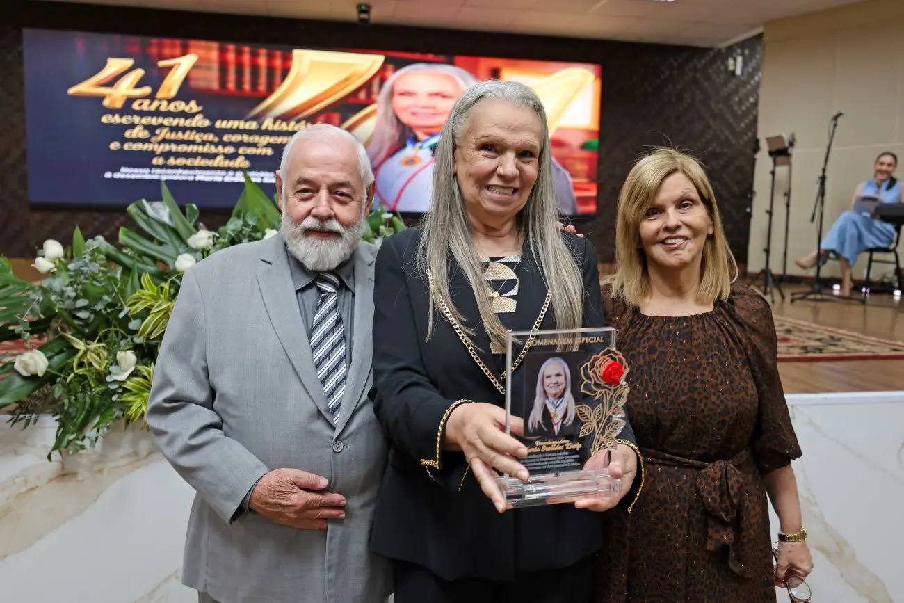 Foto horizontal que mostra a desembargadora Maria Erotides ladeada pelo presidente e pela vice-presidente do TJMT, exibindo uma placa de acrílico com sua foto e a imagem de uma rosa. Eles estão no auditório do TJMT. 