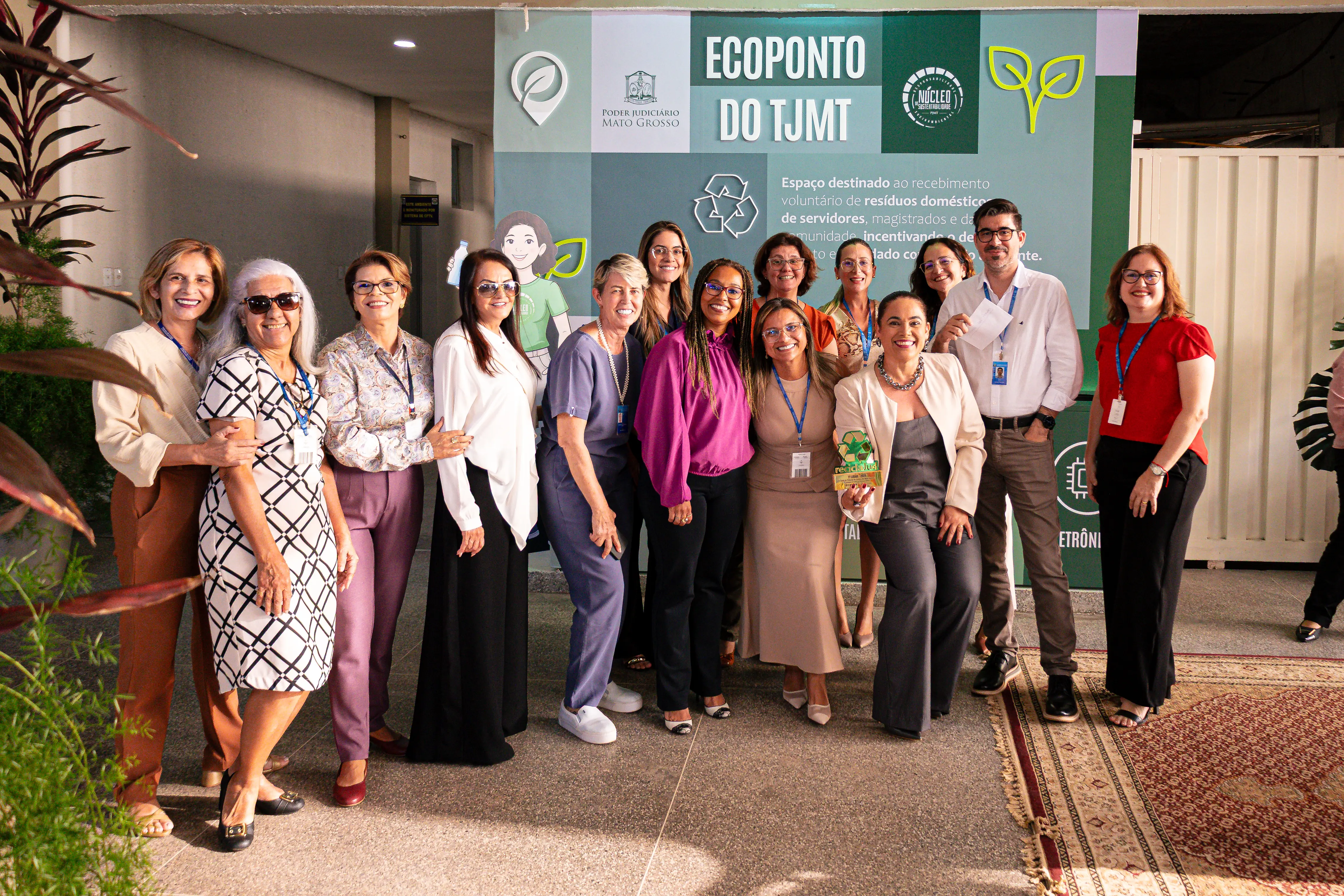 Uma fotografia posada de um grupo grande, composto por 11 mulheres e um homem, em frente ao painel do Ecoponto. O clima é de celebração e todos sorriem para a câmera. O grupo está vestido em trajes esporte fino, com roupas coloridas, terninhos, blusas sociais e vestidos.