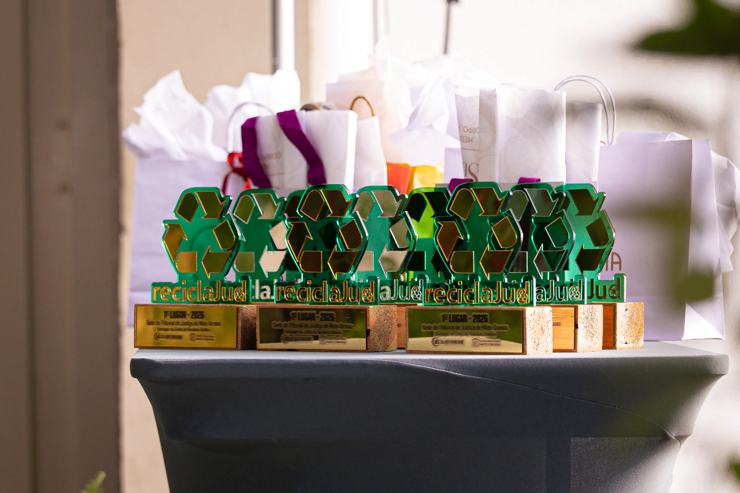 Troféus da premiação ReciclaJud, com símbolo da reciclagem em destaque, organizados sobre uma mesa. Ao fundo, sacolas de presentes entregues aos vencedores.