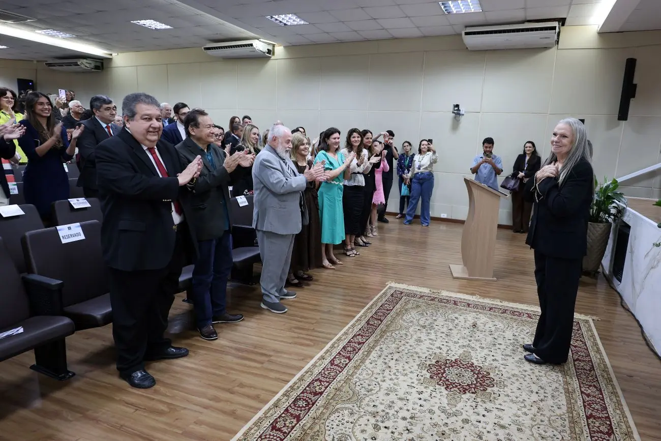 Foto horizontal em plano aberto que mostra o auditório do TJMT lotado de pessoas de pé, aplaudindo a desembargadora Maria Erotides Kneip, que está à frente, sorrindo, com as mãos sobre o peito.