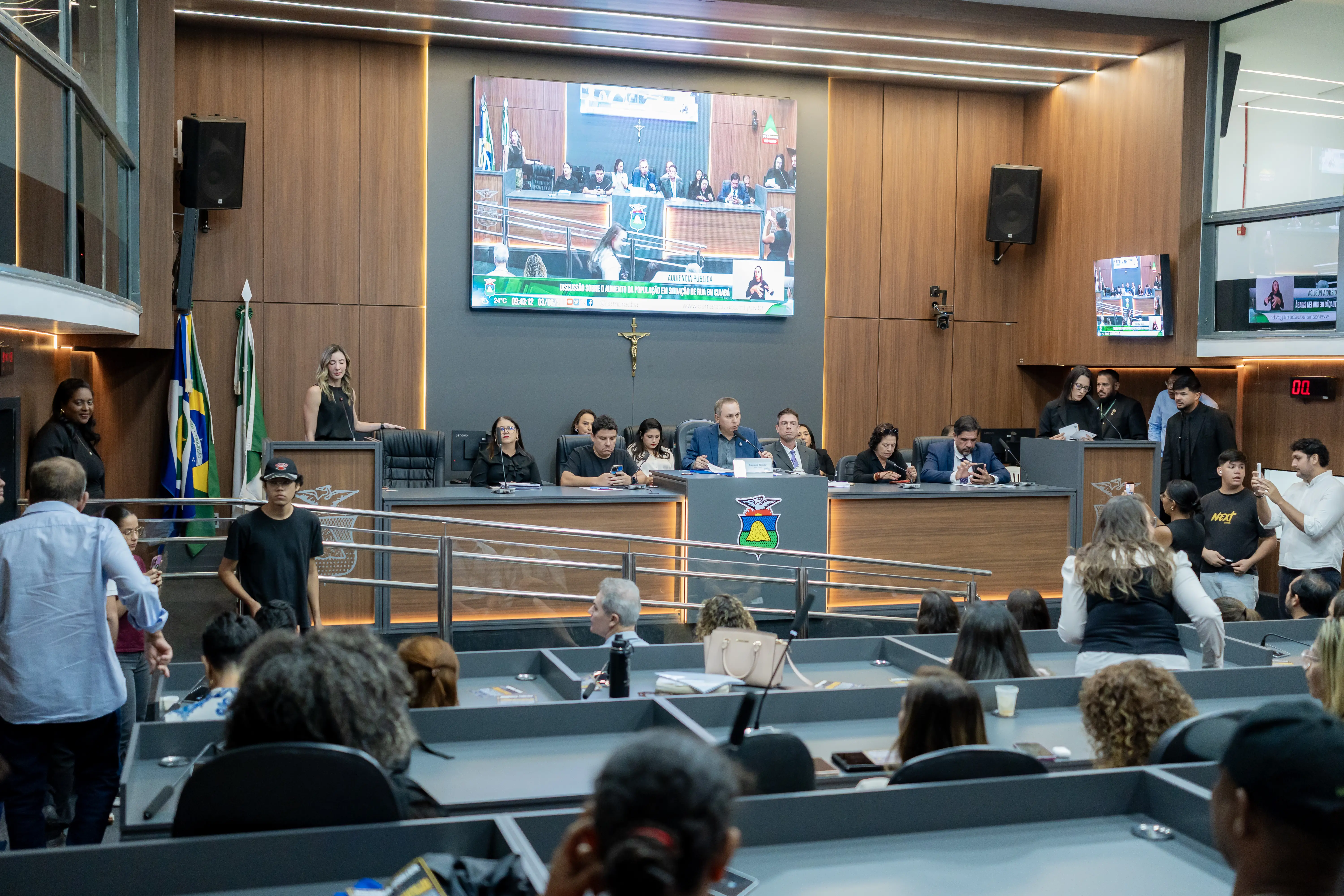 Plenário com pessoas sentadas em bancadas em primeiro plano. Ao fundo, uma mesa diretora com autoridades e um grande telão exibindo a transmissão do próprio evento.